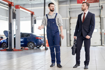 Full length portrait of modern bearded mechanic giving giving tour of workshop to handsome businessman walking across car service and repair center, copy space