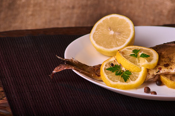 Baked fish with lemon, spices and herbs on white plate on wooden background without head