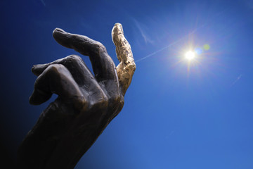 Hand of bronze statue with a finger indicating to the sky, concept collage against blue sky and sun with aureole