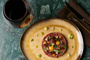 Beef tartare Steak with egg yolk, pistachios, truffle oil, green mayonnaise on porcelain plate and glass of red wine on restaurant table. Classic tartare meat with ingredients. Luxury food
