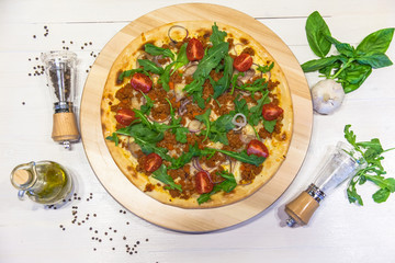 appetizing pizza with minced meat, small tomatoes, arugula spices and olive oil on a white table, flat lay