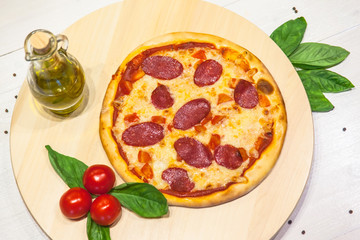 a small appetizing pizza with sausages, basil, tomatoes and olive oil on a white table
