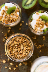 Crunchy almond and oatmeal granola in jar with yogurt kiwi granola parfait in glasses on the side, photographed overhead with natural light (Selective Focus on the top of the food)