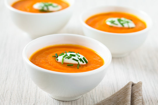 Fresh homemade cream of carrot soup with dollop of cream or yogurt and chives on top served in bowls, photographed with natural light (Selective Focus, Focus in the middle of the first soup)