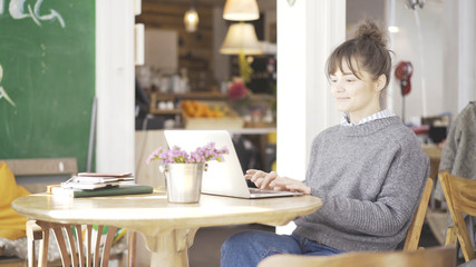 A young smiling cute happy brunette girl dressed in a grey pullover is working with a laptop in a cafe