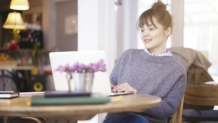 A young surprised delightful cute happy brunette girl dressed in a grey pullover is working with a laptop in a cafe