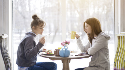 Two young adult female friends eating salad drinking tea sitting near a big window indoors in a cafe