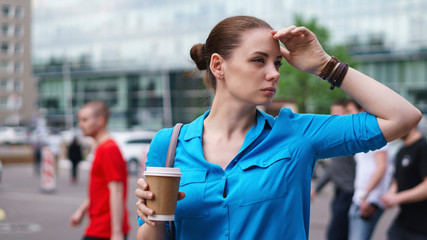 Young attractive girl is holding cup of coffee and looking away. She stands on street of city.