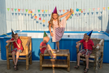 Fototapeta na wymiar Rural funny children in pointed shiny hats are playing at party near the frame swimming pool at sunset. Concept of healthy lifestyle in nature, love of peace. Girl and the boys are happy together