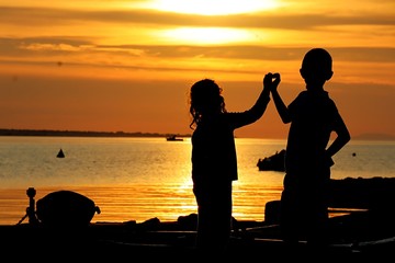 Fototapeta na wymiar Silhouettes of little boy and girl trying to make shape of heart by joining hands on marina molo during sunset 