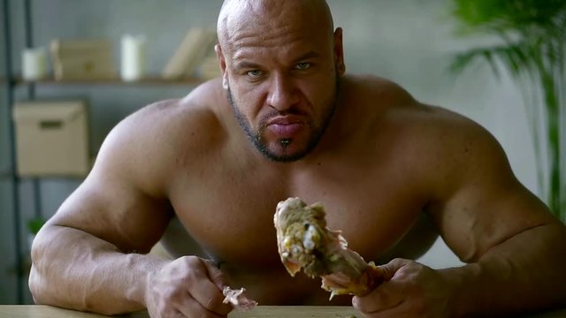 hungry brawny man with naked shoulders and chest is sitting alone in home and eating fried leg, biting meat