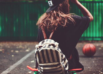 Young girl sitting on plastic orange penny shortboard on asphalt in cap