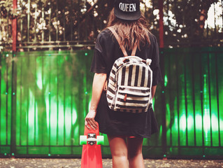 Young girl with plastic orange penny shortboard behind green wall in cap