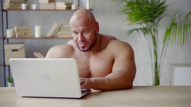 brutal naked man is typing on keyboard of notebook, writing text on digital gadget, sitting alone in room