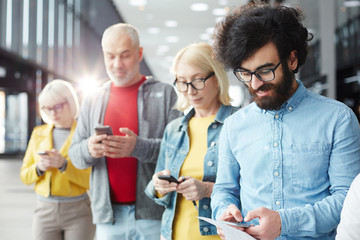 Row of people in casualwear searching in their smartphones while waiting for their turn
