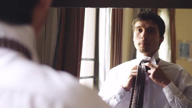 Young man in white shirt stands by the mirror tying a tie. 3840x2160