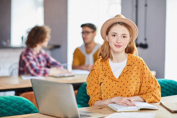 Young businesswoman sitting by workplace, making working notes and networking with colleagues talking on background