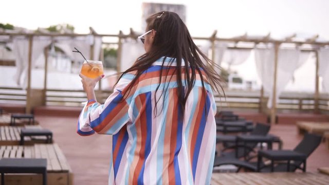 Traking footage of a luxury, fashion woman with a glass of cocktail walking colored silk dressing gown by the wooden lounge area near the sea. Summertime