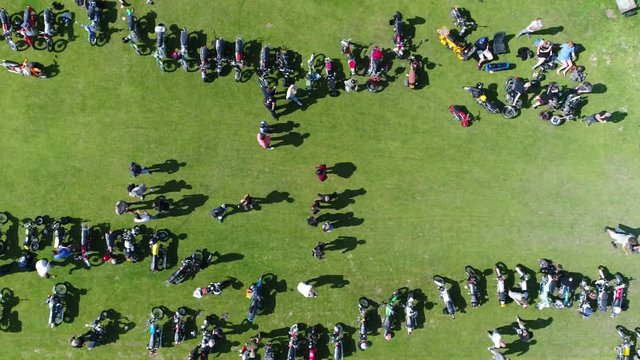 Aerial top down footage of moped event moving right over green field with the small motorcycles parked and people walking over the festival terrain steady footage during summer day 4k high resolution