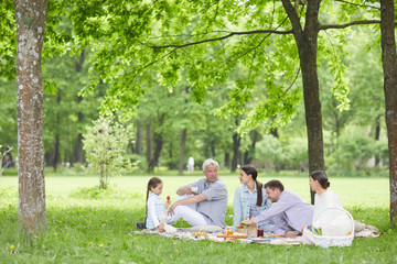 Two couples and cute little girl enjoying picnic on green lawn on hot summer day