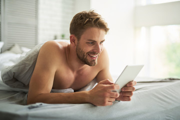 Side view of carefree smiling male lying on belly in bed with tablet in hands. He is looking at screen with pleasure and delight