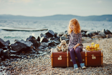 A little sad girl with a toy sitting on a large suitcase near th