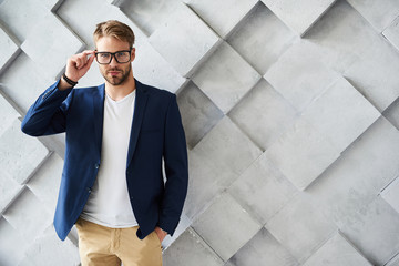 Waist up portrait of handsome stylish man standing and touching the rim of spectacles. He is keeping his hand in pocket while looking straightforward with assurance. Copy space in right side