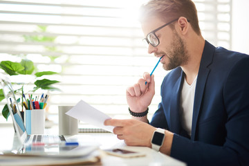 Profile of businessman concentrated on checking documentation of company. He is sitting at desktop...