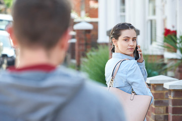 Young Woman Calling For Help On Mobile Phone Whilst Being Stalked On City Street By Man - obrazy, fototapety, plakaty