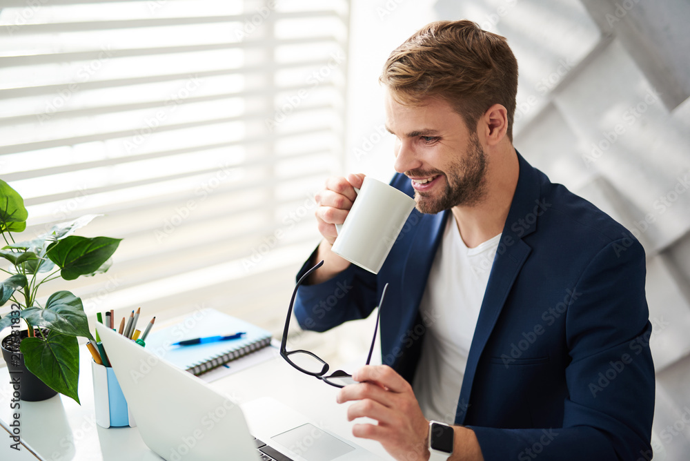 Poster side view of smiling man drinking favorite hot drink in office with eyeglasses in hands. he is sitti