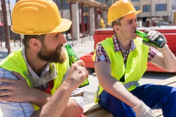Poster Two young workers smiling, while drinking a cold alcoholic or non-alcoholic beer during break at work on the construction site in a sunny day of summer © Kzenon