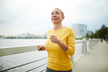 Mature active blond woman in yellow pullover jogging on city bridge along riverside in the morning