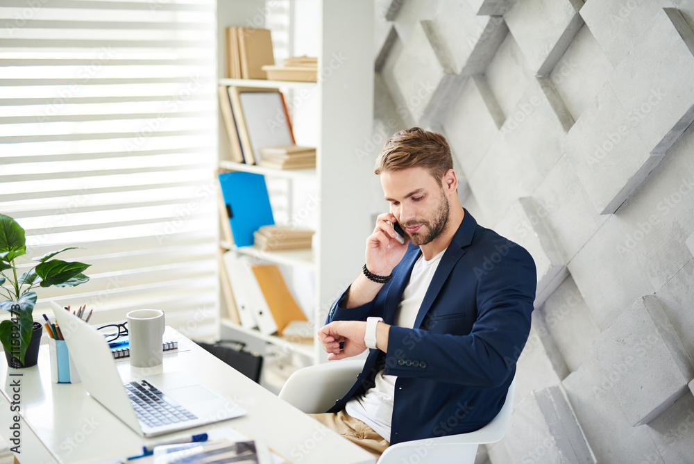 Poster side view of man sitting at laptop and checking time. he is talking on phone and discussing business
