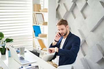 Naklejka premium Side view of man sitting at laptop and checking time. He is talking on phone and discussing business in relaxed way