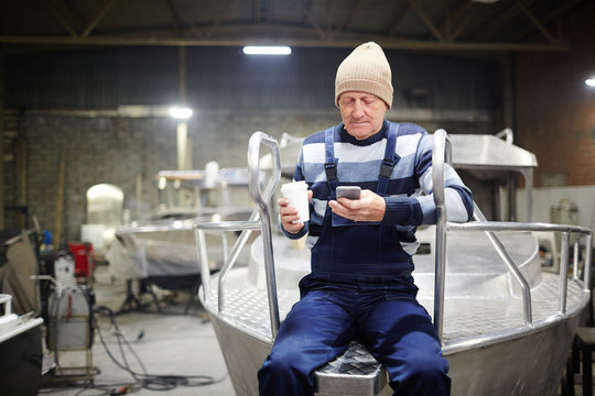 Aged Engineer With Drink And Smartphone Messaging While Sitting On Motorboat In Workshop