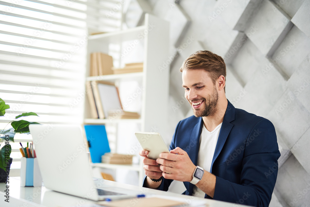 Poster side view of happy male sitting at table with computer on it. he is using portative device for fun w