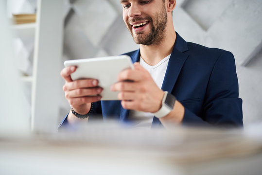 Low angle of male smiling at work. Businessman is sitting at table and using tablet with joy and content