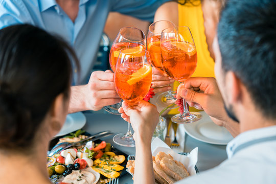 Cheerful young best friends toasting with a refreshing delicious summer drink while sitting together at table at a trendy restaurant outdoors