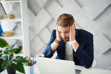Oh my God. Confused businessman working on project at computer. He is holding head with hand and...