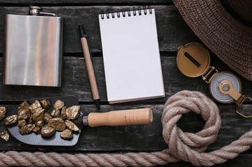 Blank notepad page, hat, flask, shovel full of gold ore, compass, rope and magnifying glass on aged wooden table background. Treasure hunter concept. Adventurer equipment.
