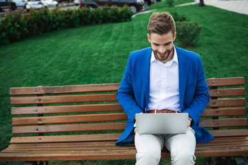 Fototapeta premium Cheerful young businessman is using laptop and laughing. He is sitting on bench in park. Copy space 