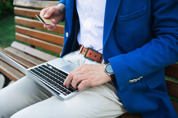 Close up of male arm doing online shopping while holding laptop. Man is sitting on bench outdoor