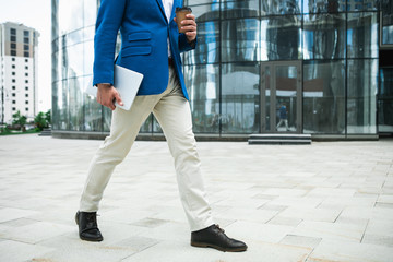 Low angle close up of male body. Busy businessman walking towards the city street. He is carrying cup of coffee and laptop. Copy space 