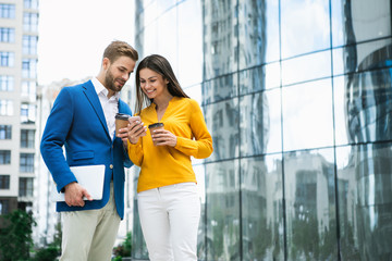 Portrait of joyful young woman is showing her mobile phone to colleague. They are standing outdoor and laughing. Friends are drinking coffee after work. Copy space 