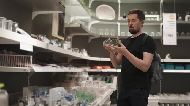 a young and bearded man chooses glass goblets in a shopping boutique, maybe he wants to equip his new apartment with comfort, so he chooses a stylish dish