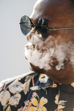 Black stylish man wearing sunglasses. He is vaping with an electronic cigarrete outdoor