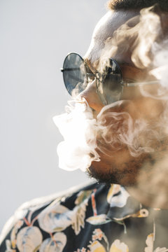 Black stylish man wearing sunglasses. He is vaping with an electronic cigarrete outdoor