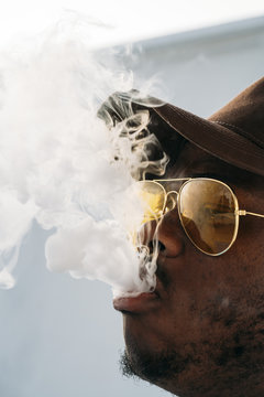 Black stylish man wearing sunglasses. He is vaping with an electronic cigarrete outdoor