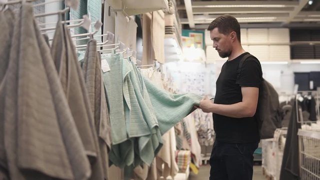 a young man who is in the mall examines on the shelves of goods for the house, Jeltelman touches a terry bath towel, he wants to know the composition of the goods