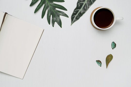 Cup of coffee and notebook on white wooden background with tropical monstera leaves. Minimal flat lay, top view.